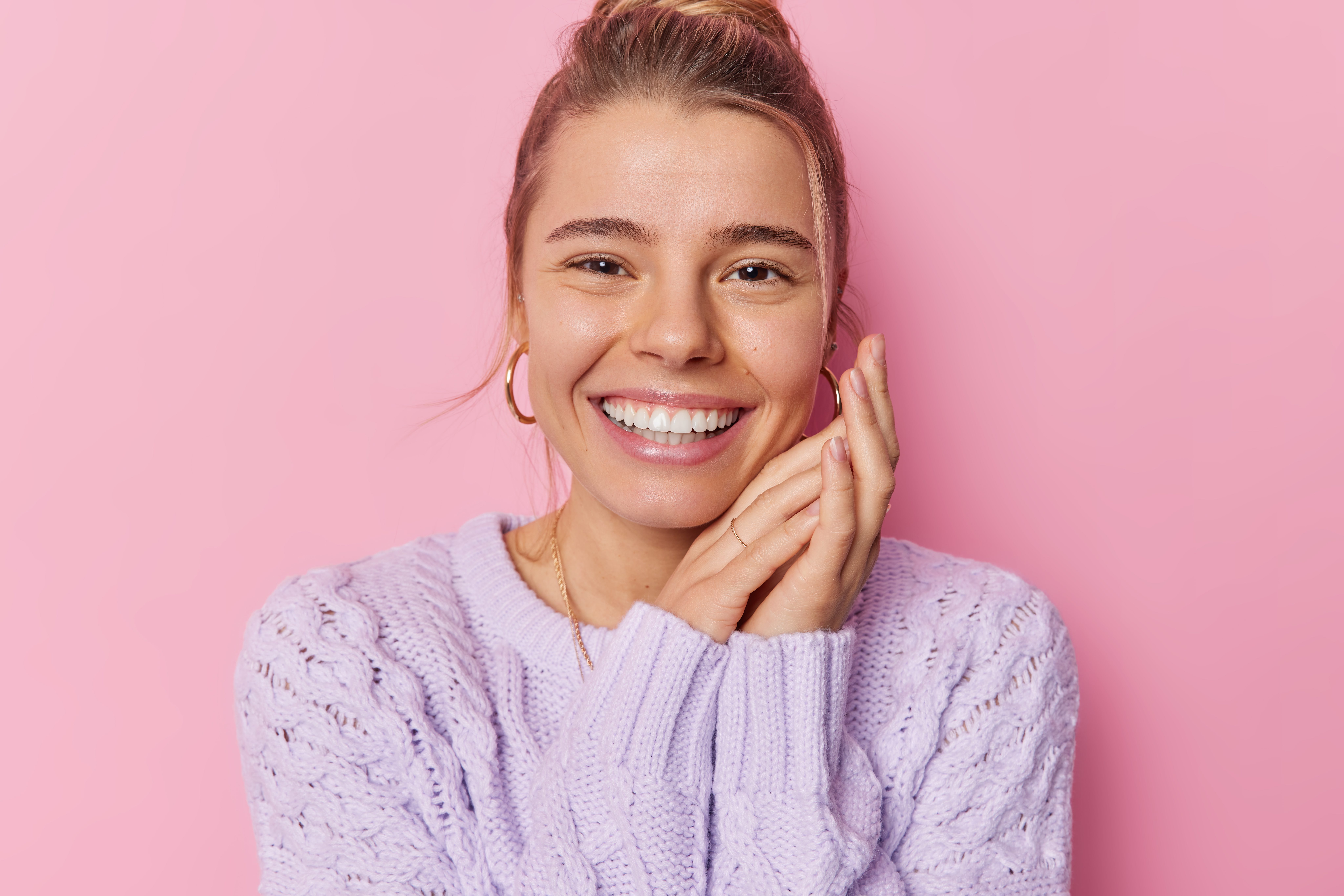 portrait-lovely-young-woman-smiles-toothily-keeps-hands-near-face-looks-glad-wears-purple-knitted-sweater-earrings-poses-against-pink-background-happy-emotions-face-expressions-concept